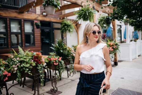 Bows & Sequins wearing a one-shoulder crop top and Call It Spring white sunglasses with a Chanel bracelet via Switch.