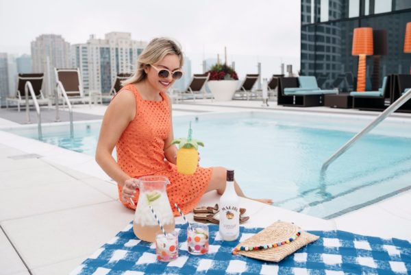 Bows & Sequins wearing an orange eyelet dress on a blue gingham picnic blanket, drinking Malibu Rum coconut cocktails, by the pool.
