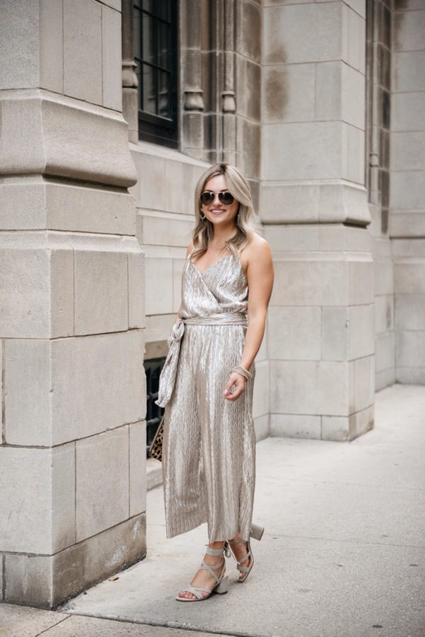 Jessica of Bows & Sequins, a fashion-focused lifestyle blog, wearing a Nordstrom tie-waist metallic gold jumpsuit with grey suede block heel sandals, Gucci sunglasses, a Clare V leopard clutch, and pavé bracelets.