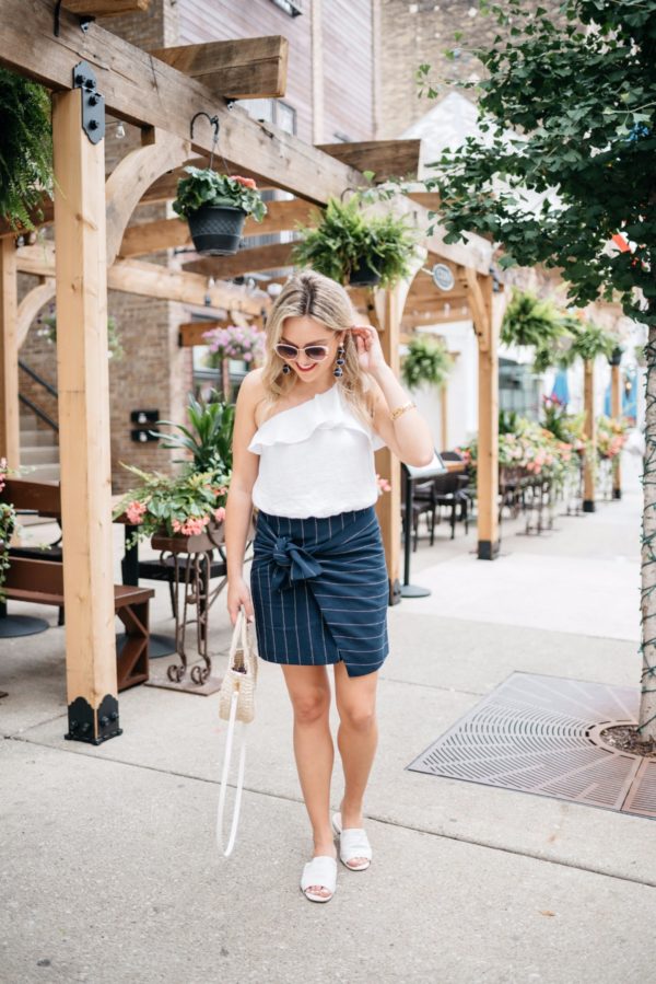 Bows & Sequins wearing a ruffle one-shoulder crop top with a tie-front pinstripe skirt, white leather slides, a straw tote, and white sunglasses.