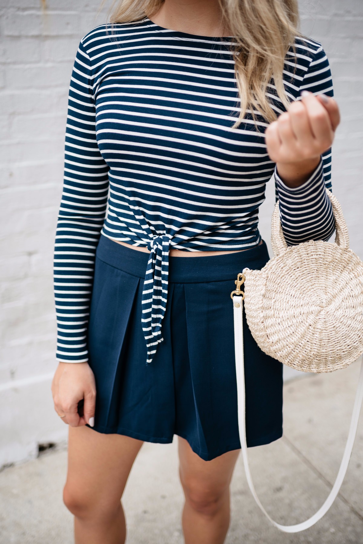 Bows & Sequins wearing a navy striped tie-front crop top with navy pleated shorts and a straw tote.
