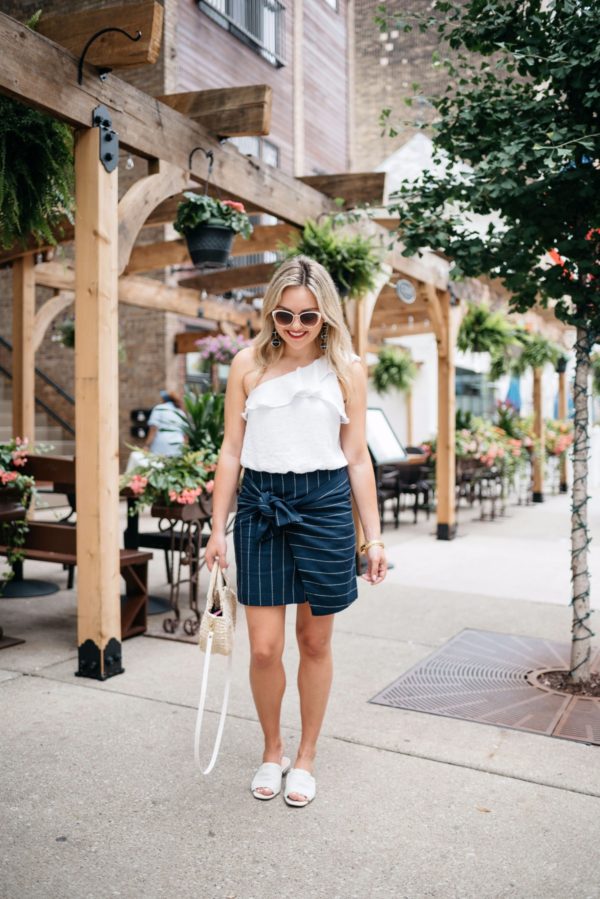 Fashion and lifestyle blogger Bows & Sequins wearing a tie-front navy pinstripe J.O.A. skirt with a one-shoulder crop top, Call It Spring sunglasses, a Clare V straw tote, and Kurt Geiger slides.