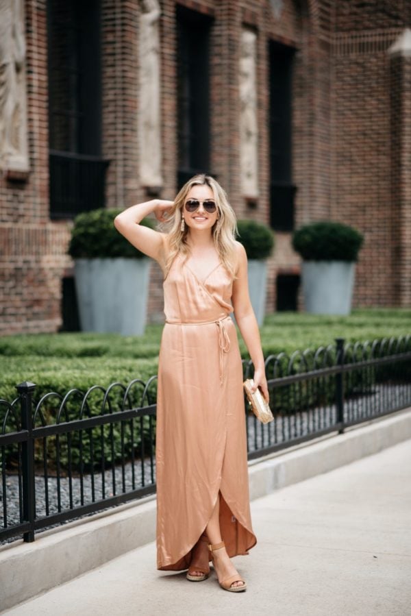 Lifestyle and fashion blogger Bows & Sequins wearing a metallic silk dress with gold aviators, a gold pouch, nude lipstick, and camel wedges.
