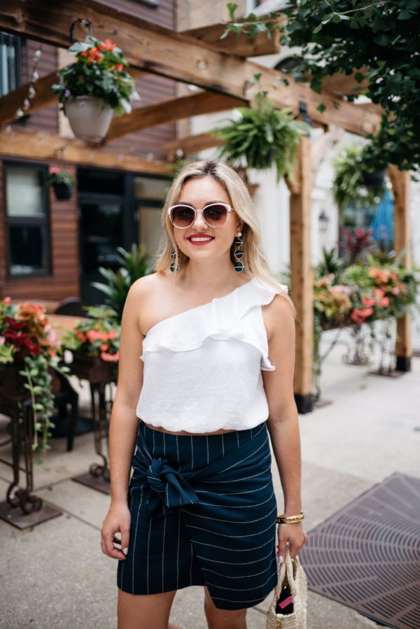 Bows & Sequins wearing Call It Spring sunglasses, an ASTR ruffle one-shoulder crop top, and a front-tie pinstripe skirt.