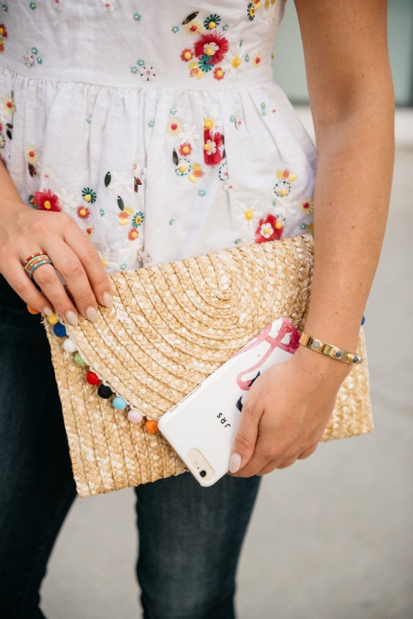 Style blogger Bows & Sequins wearing a J.Crew embroidered top and a BaubleBar cuff, holding a White Elephant Designs pom pom straw clutch and a Minnie & Emma flamingo phone case.