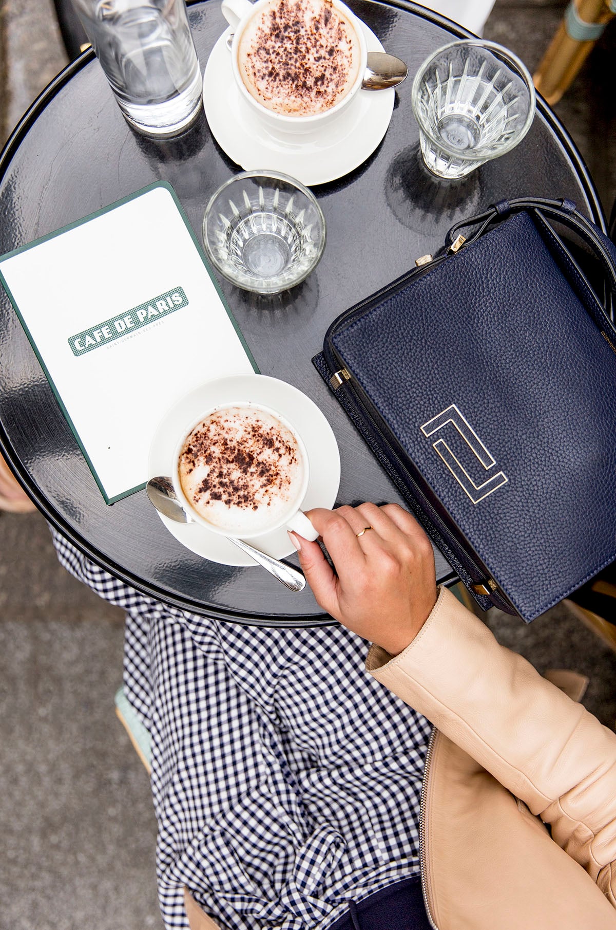 Bows & Sequins wearing gingham pants and a navy leather Lancel Pia Bag drinking cappuccinos at Cafe de Paris in Saint Germain des Pres.
