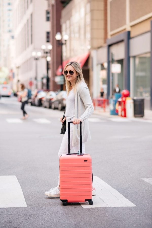 Bows & Sequins wearing an all white outfit with a pink hard shell carry on suitcase.