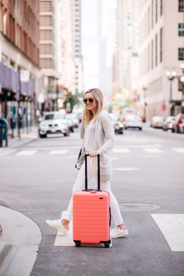 Bows & Sequins wearing an all white outfit with a pink hard shell carry on suitcase.