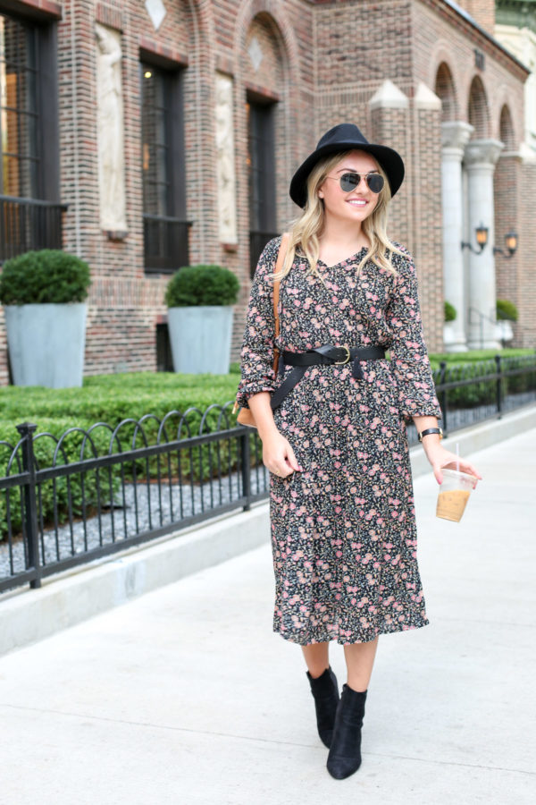 Jessica of the fashion-focused blog Bows & Sequins, wearing a black Old Navy hat, matte aviators, a black leather Madewell belt, and ankle booties with her Old Navy fall floral dress.