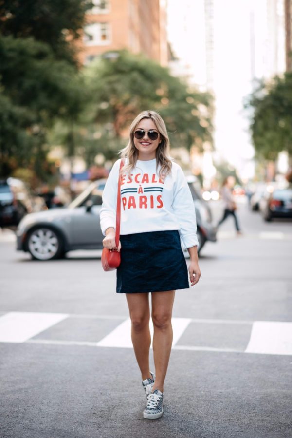 Fashion and travel writer Bows & Sequins wearing a French Disorder sweatshirt with an Old Navy blue suede skirt, a Talbots red leather bag, aviators, and Claudie Pierlot gingham sneakers.