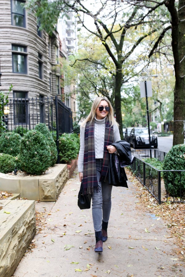 Jessica Sturdy of Bows & Sequins wearing a Barbour black Beadnell jacket with a cable knit grey sweater, grey jeans, and patchwork booties with a tartan scarf in Chicago.