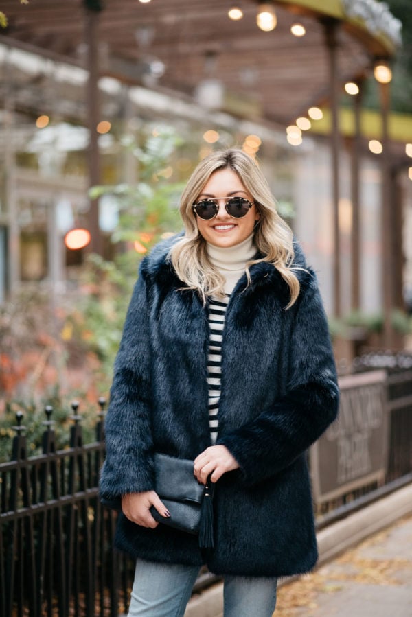 Jessica Sturdy wearing a Miranda Dunn London faux fur coat with a striped turtleneck sweater, Illesteva sunglasses, and an Azalea Blue foldover clutch.