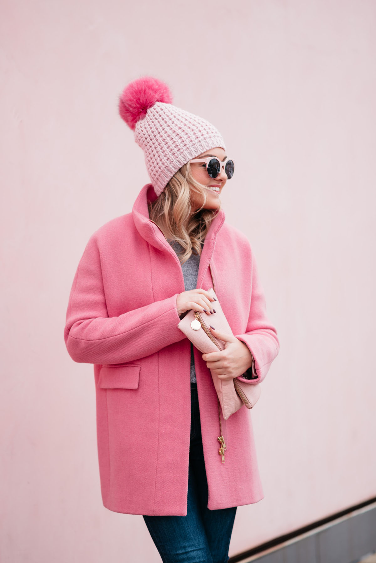 Fashion blogger Jessica Rose Sturdy wearing pink sunglasses, a pink pom pom hat, a blush pink clutch, and a J.Crew cocoon coat.