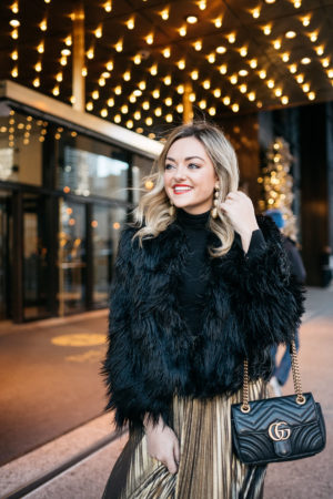 Jessica Rose Sturdy wearing a black and gold holiday outfit: 1.State black faux fur jacket, black turtleneck, Eliza J gold pleated maxi skirt, Kenneth Jay Lane gold ball earrings and Dolce & Gabbana red lipstick with a Gucci Marmont bag at the Langham in Chicago.