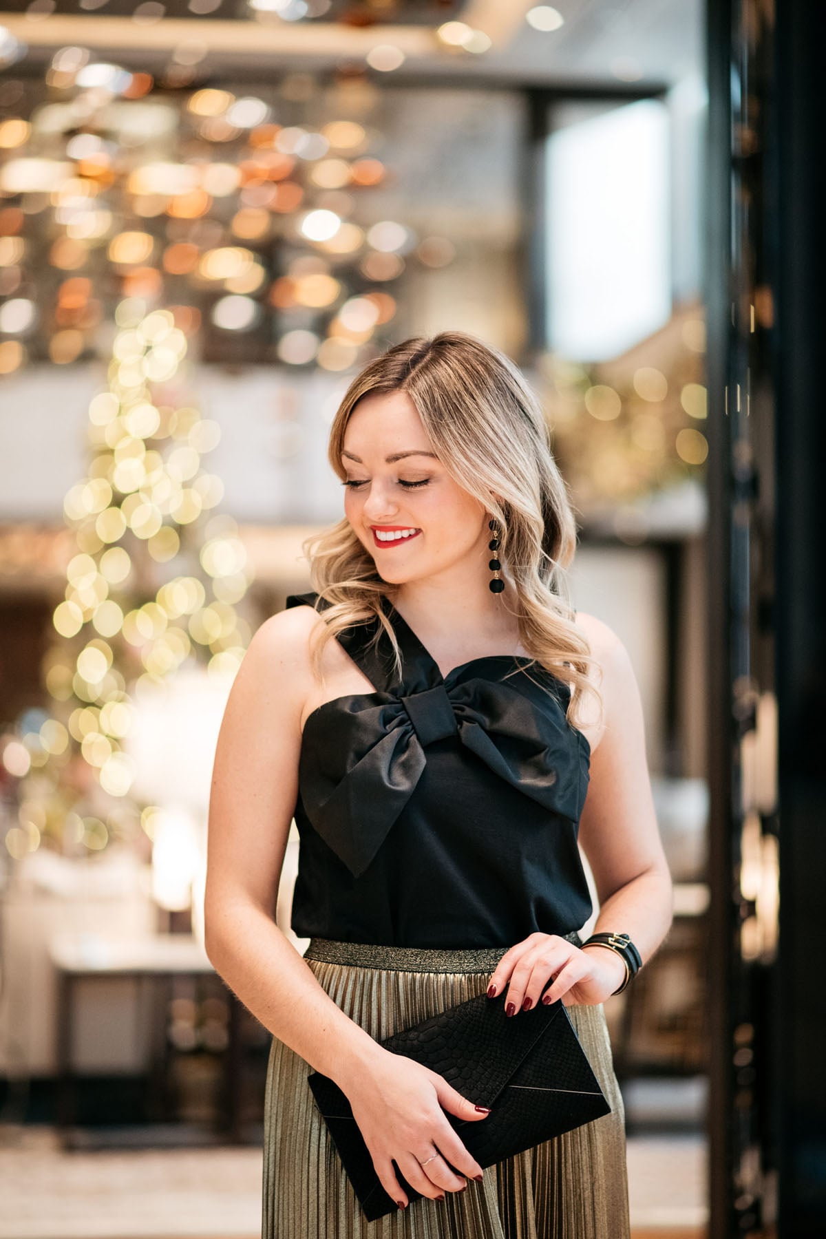 Jessica Sturdy wearing Baublebar black ball earrings, a black and gold cuff, and J.Crew black bow top with a gold pleated skirt for the holidays.