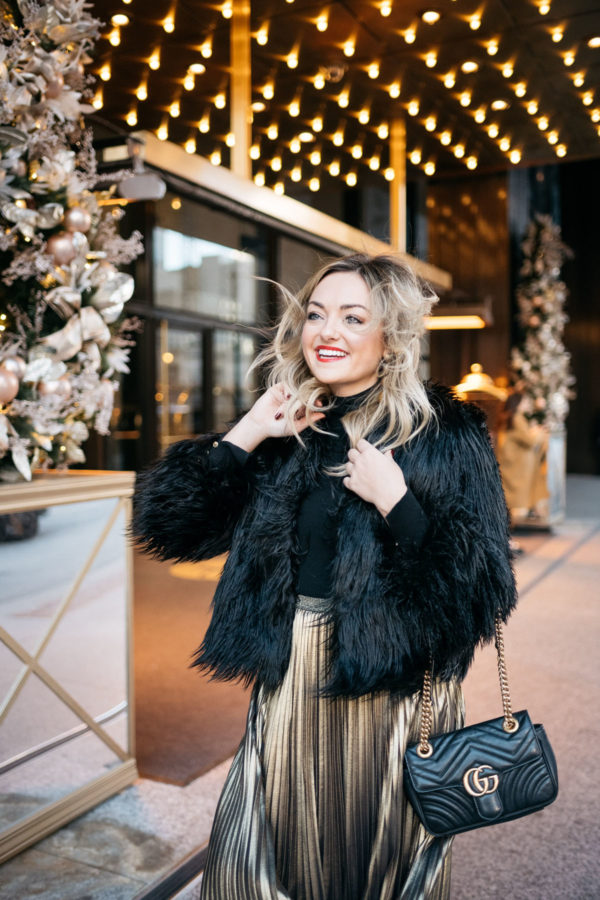 Jessica Rose Sturdy wearing a black and gold holiday outfit: 1.State black faux fur jacket, black turtleneck, Eliza J gold pleated maxi skirt, Kenneth Jay Lane gold ball earrings and Dolce & Gabbana red lipstick with a Gucci Marmont bag at the Langham in Chicago.