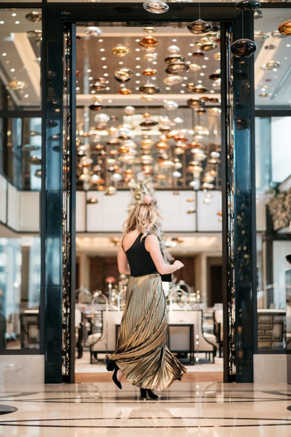 Chicago lifestyle blogger Jessica Rose Sturdy wearing a J.Crew black bow top, Eliza J gold pleated maxi skirt, Marc Fisher black velvet pumps, and a Baublebar black and gold cuff with a black envelope clutch at the Langham.