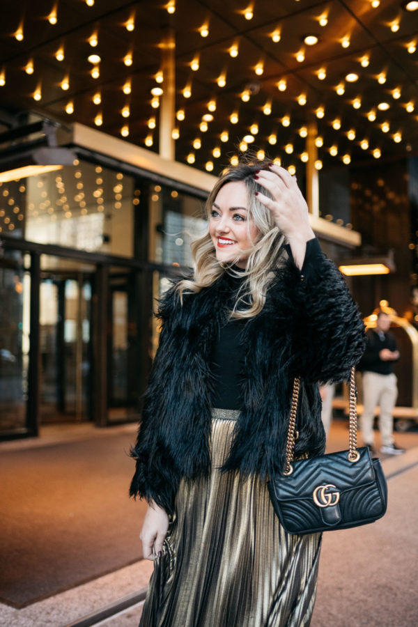 Jessica Rose Sturdy wearing a black and gold holiday outfit: 1.State black faux fur jacket, black turtleneck, Eliza J gold pleated maxi skirt, Kenneth Jay Lane gold ball earrings and Dolce & Gabbana red lipstick with a Gucci Marmont bag at the Langham in Chicago.