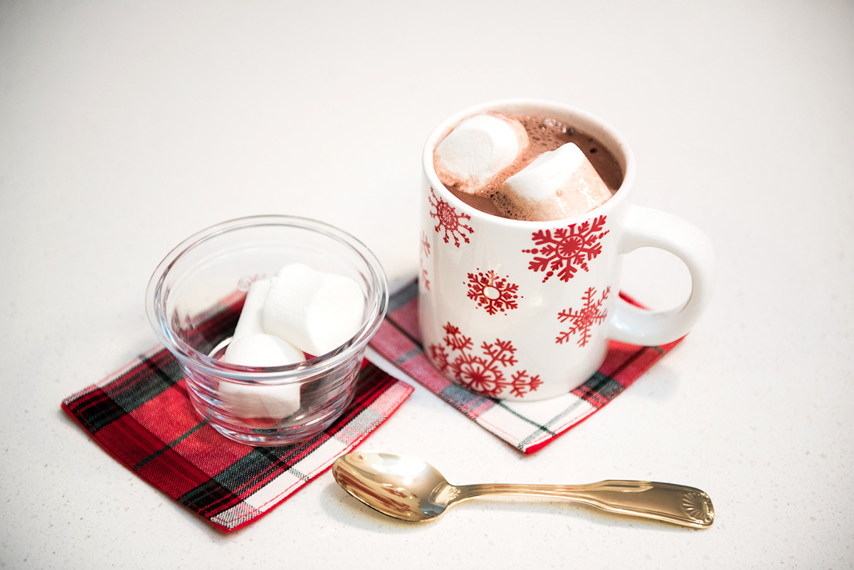 Hot chocolate with marshmallows in a snowflake mug with tartan plaid coasters and a gold spoon.