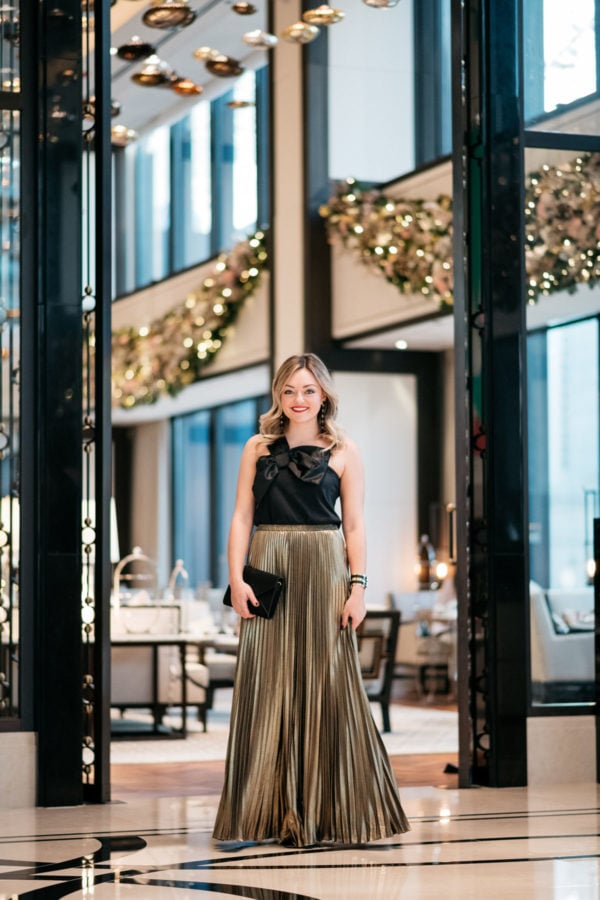 Jessica Rose Sturdy wearing a J.Crew black bow top, Eliza J gold pleated maxi skirt, Marc Fisher black velvet pumps, and a Baublebar black and gold cuff with a black envelope clutch at the Langham in Chicago.