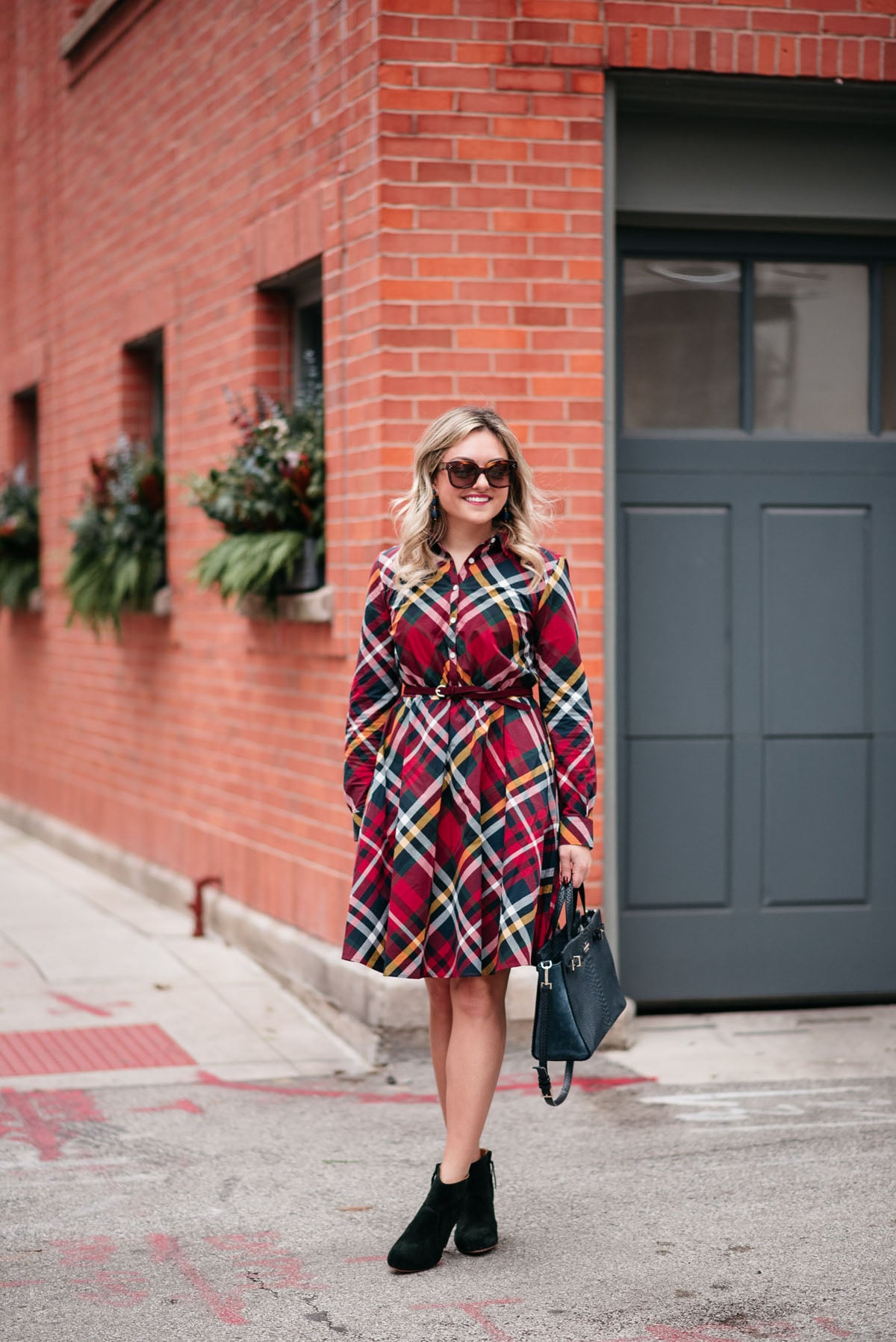 Jessica Rose Sturdy wearing tortoise sunglasses, a Brooks Brothers plaid dress, and booties with statement earrings and a navy Kate Spade tote.