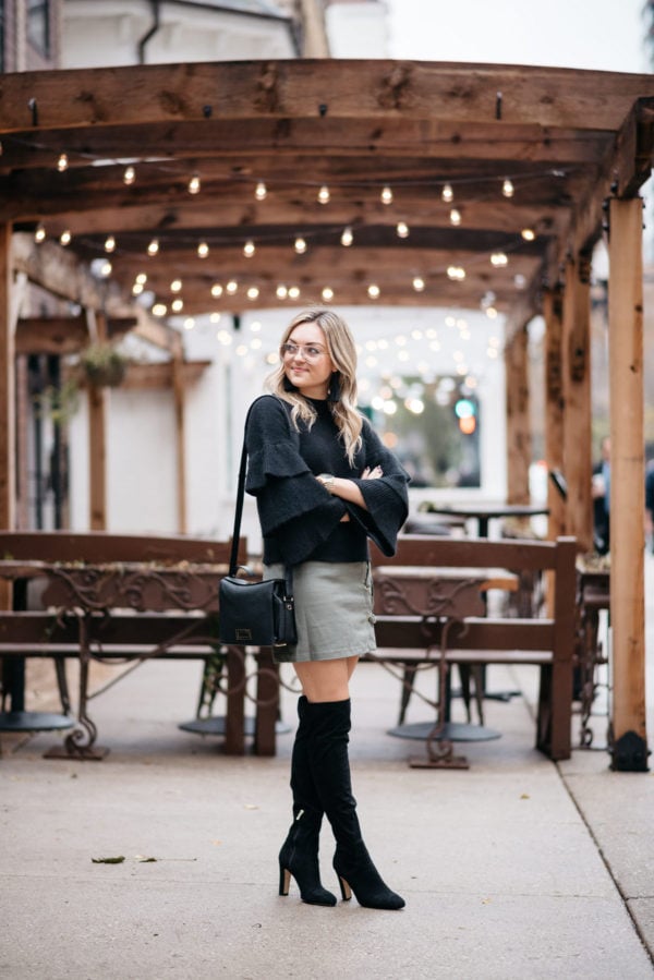 Jessica Sturdy wearing a black Endless Rose ruffle sleeve sweater, a suede lace-up skirt, black over the knee boots, and H&M clear glasses with a Lancel crossbody bag and Michael Kors watch.