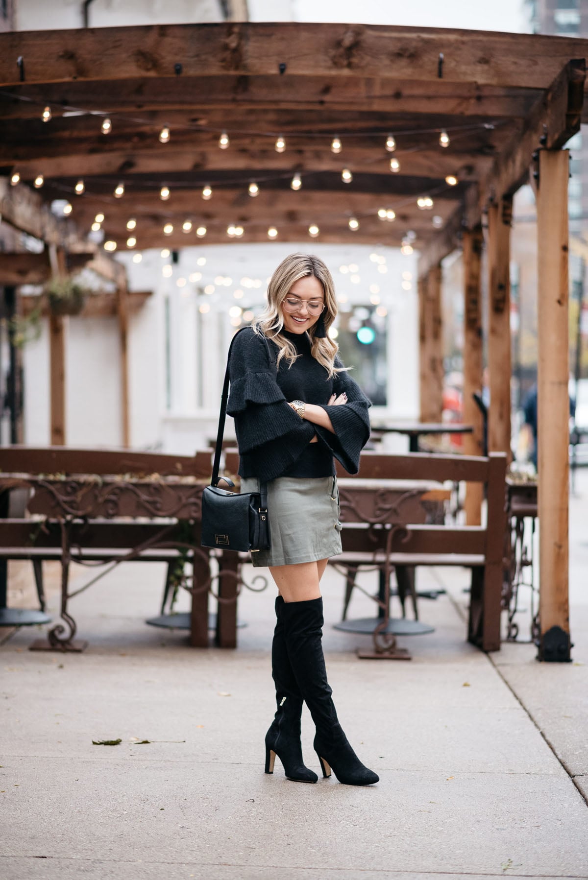Jessica Sturdy wearing an Endless Rose black ruffle sleeve sweater, a Kensie suede lace-up skirt, black over the knee boots, and H&M clear glasses with a Lancel crossbody bag, Hart tassel earrings, and Michael Kors watch.