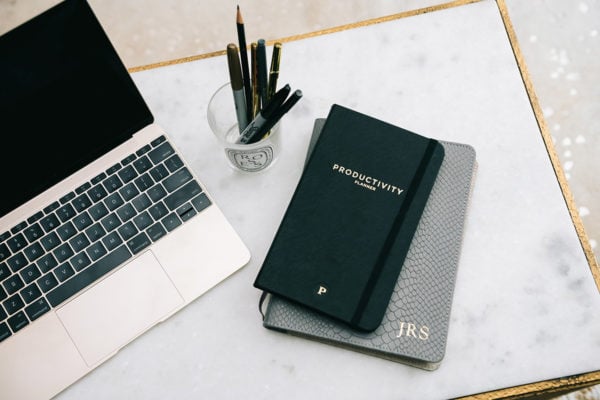 A gold MacBook laptop on a marble table with a Diptyque jar of markers, pens, and pencils, alongside a Productivity Planner and a monogrammed leather Journal.