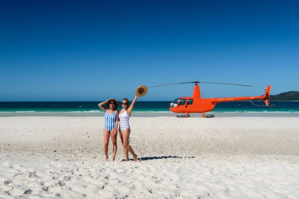 Hallie Wilson and Jessica Sturdy wearing striped one piece swimsuits in front of an orange helicopter on Whitehaven Beach in the Whitsundays in Australia