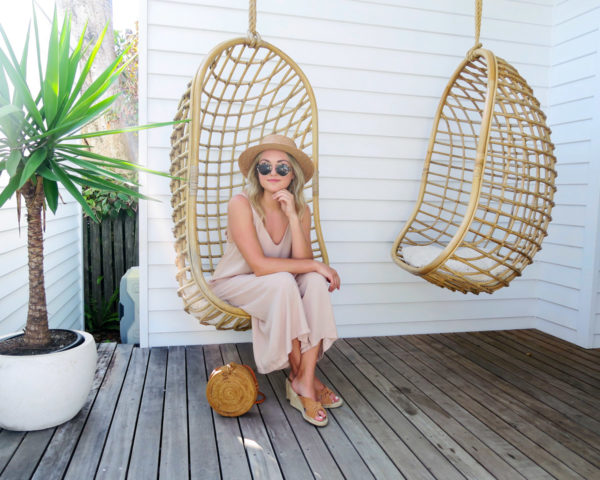 Jessica Sturdy wearing a jumpsuit and a straw hat in a swinging rattan chair at Bask & Stow in Byron Bay in Australia