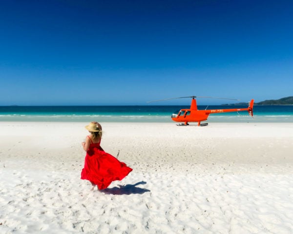 Jessica Sturdy on Whitehaven Beach in the Whitsundays in Australia wearing a red maxi dress in front of an orange helicopter.