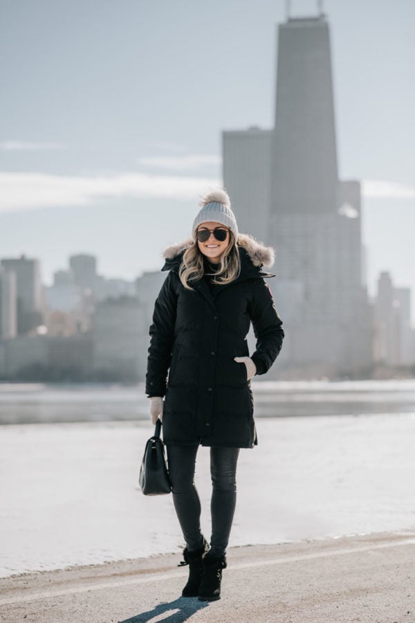 Jessica Sturdy wearing a fur-trimmed, hooded Canada Goose jacket with a pom beanie, matte aviators, booties, and a Polene top handle bag.
