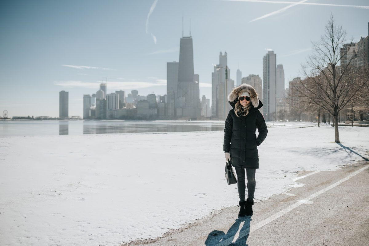 Jessica Sturdy wearing a fur-trimmed, hooded Canada Goose jacket with a pom beanie, matte aviators, booties, and a Polene top handle bag by Lake Michigan.