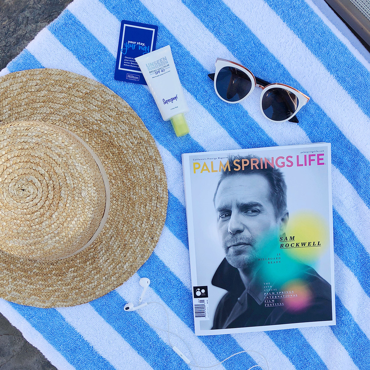 Jessica Sturdy shares a poolside photo from the Miramonte Resort in Indian Wells with a Palm Springs Life Magazine, blue striped towel, L*Space straw hat, Supergoop Unseen sunscreen, and white sunglasses.