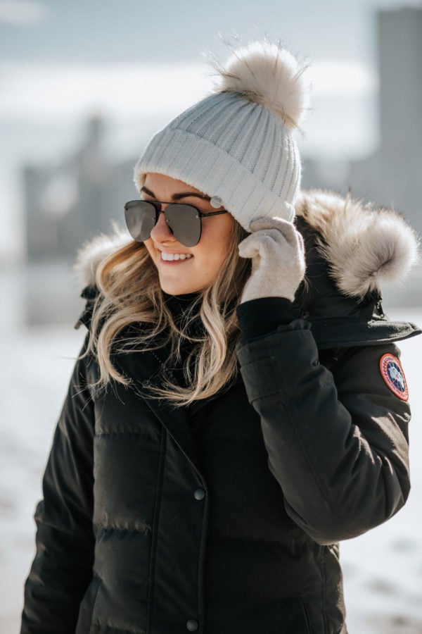 Jessica Sturdy wearing a fur-trimmed, hooded Canada Goose jacket with a pom beanie and matte aviators.