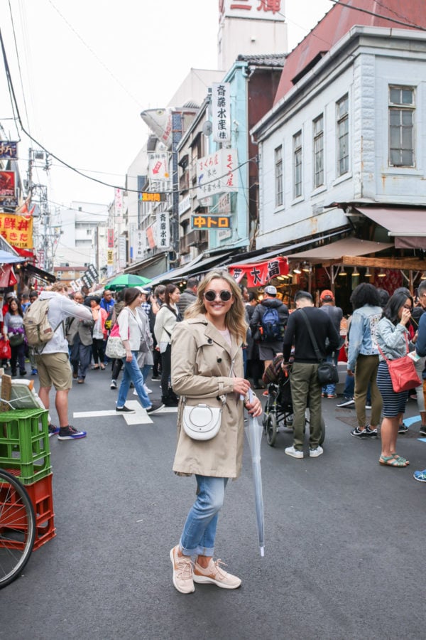 Travel blogger Jessica Sturdy at Tsukiji Market in Tokyo, Japan wearing a tan trench coat, white Theory crossbody ring bag, jeans, sneakers, and a clear umbrella.