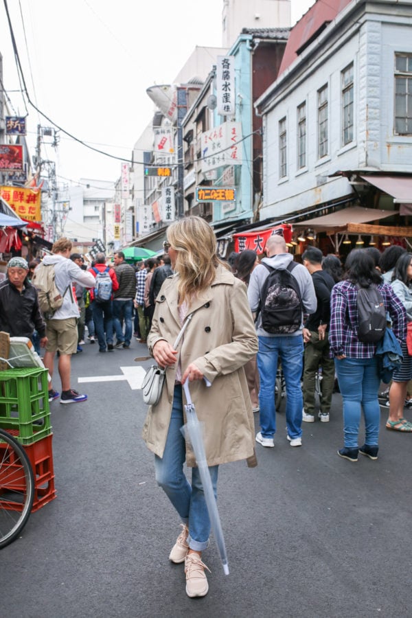 Travel blogger Jessica Sturdy at the Tsukiji Market in Tokyo Japan wearing a trench coat, cuffed jeans, and sneakers with a clear umbrella on a rainy day.