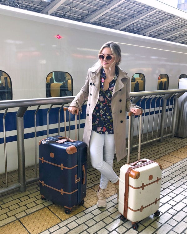 Bows & Sequins Travel Guide in Tokyo Japan. Jessica Sturdy is wearing a trench coat, Rebecca Taylor floral blouse, white jeans, Timberland blush pink sneakers, pink sunglasses, and two Brics Luggage suitcases at the JR Shinkansen platform at Tokyo Station