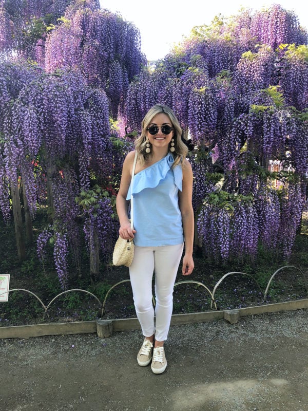 Bows & Sequins Travel Guide in Ashikaga Flower Park in Japan. Jessica Sturdy by purple wisteria flowers wearing a Vineyard Vines ruffled one-shoulder top, Old Navy white jeans, Seavees raffia sneakers, and a Clare V straw bag.