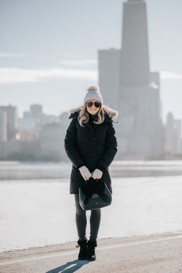 Jessica Sturdy wearing a fur-trimmed, hooded Canada Goose jacket with a pom beanie, matte aviators, booties, and a Polene top handle bag.