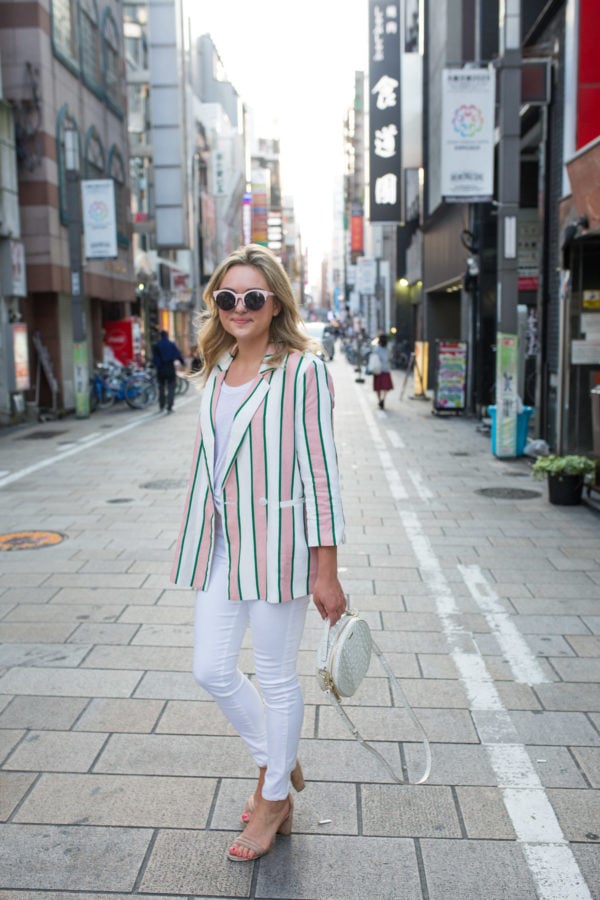 Fashion blogger Jessica Sturdy styling white jeans with a striped blazer, pink sunglasses, and white Brahmin bag in Osaka, Japan.