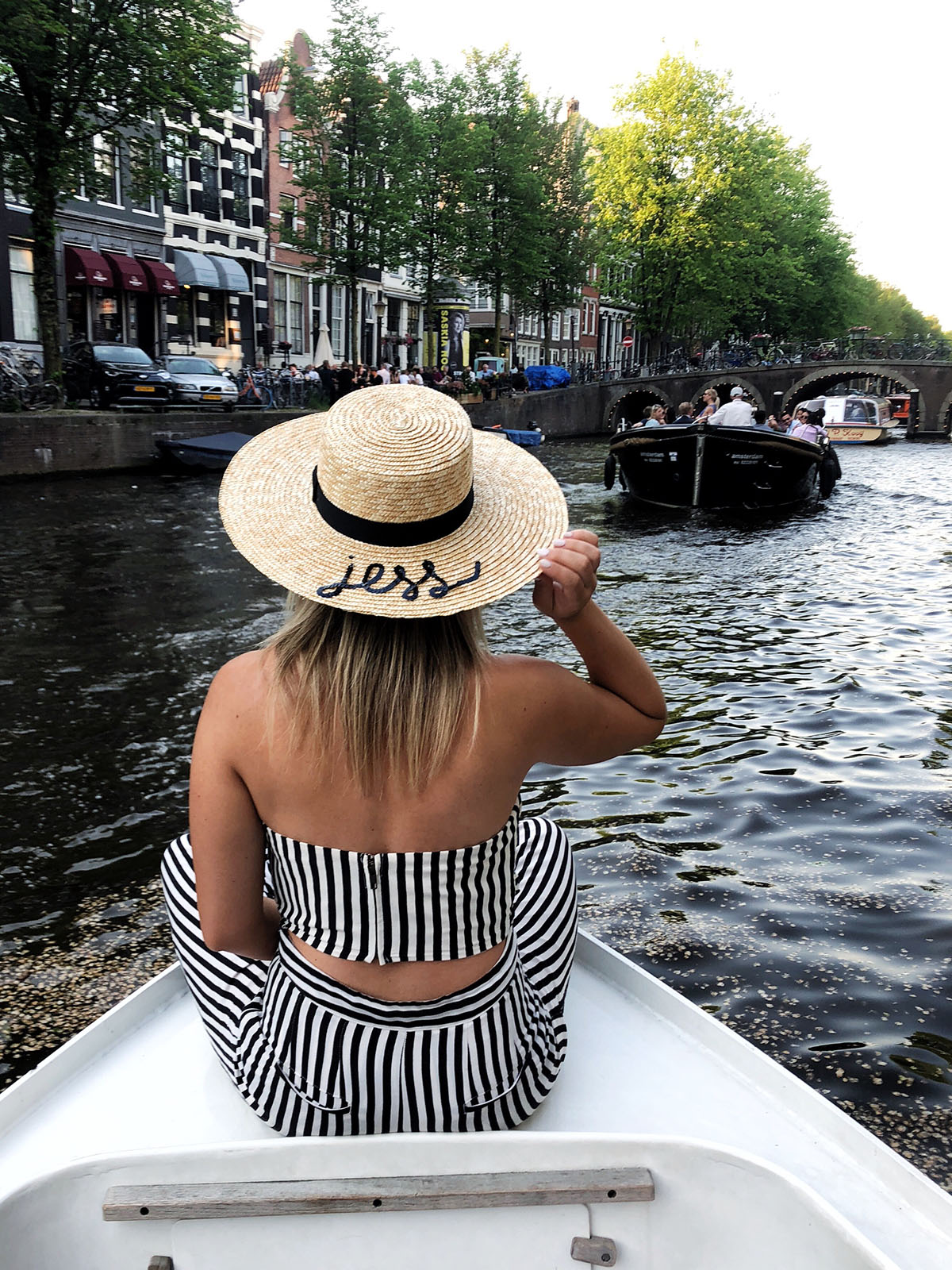 Jessica Sturdy wearing a black and white striped Lovers + Friends crop top and pants set with a monogramed straw hat that says Jess on the front of a boat in the scenic canals of Amsterdam.