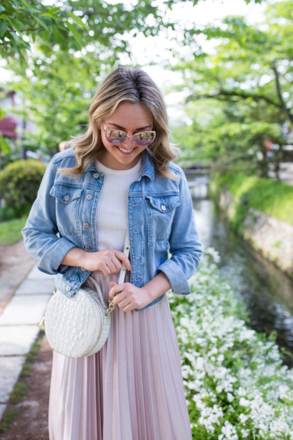 Chicago travel-based blogger and influencer Jessica Sturdy styling a denim jacket with a tee, pleated skirt, and round handbag in Kyoto, Japan along the Philosopher's Path walkway.