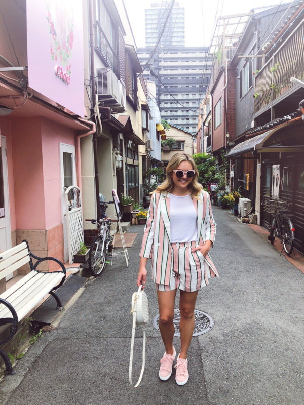 Jessica Sturdy wearing a white and pink striped blazer and short suit set, pink Vanessa Wu sneakers, pink sunglasses, and a round Brahmin white bag in the hipster neighborhood of Nakazakicho in Osaka, Japan.