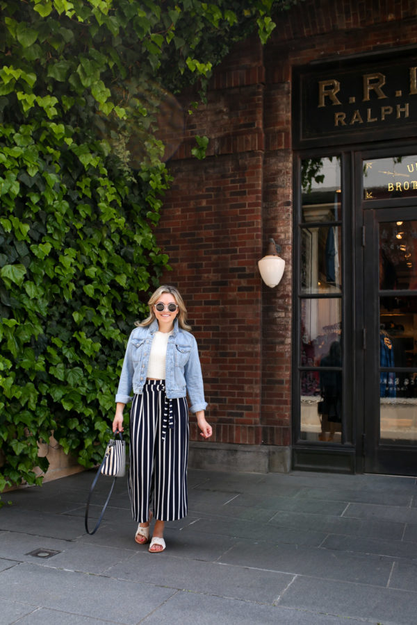 Fashion blogger Jessica Sturdy wearing a jean jacket, crop top, and striped wide leg pants in front of a green ivy and red brick wall in Tokyo, Japan.