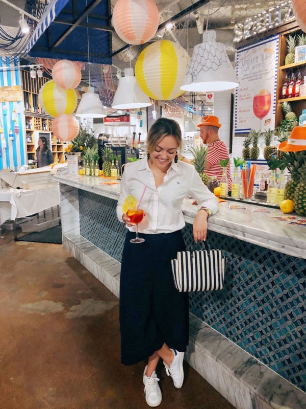 Jessica Sturdy at the Eataly Chicago Sabbia opening with an Aperol Spritz wearing a white button front shit, wide leg pants, Adidas stan smith sneakers, and a stripe Clare V bag.