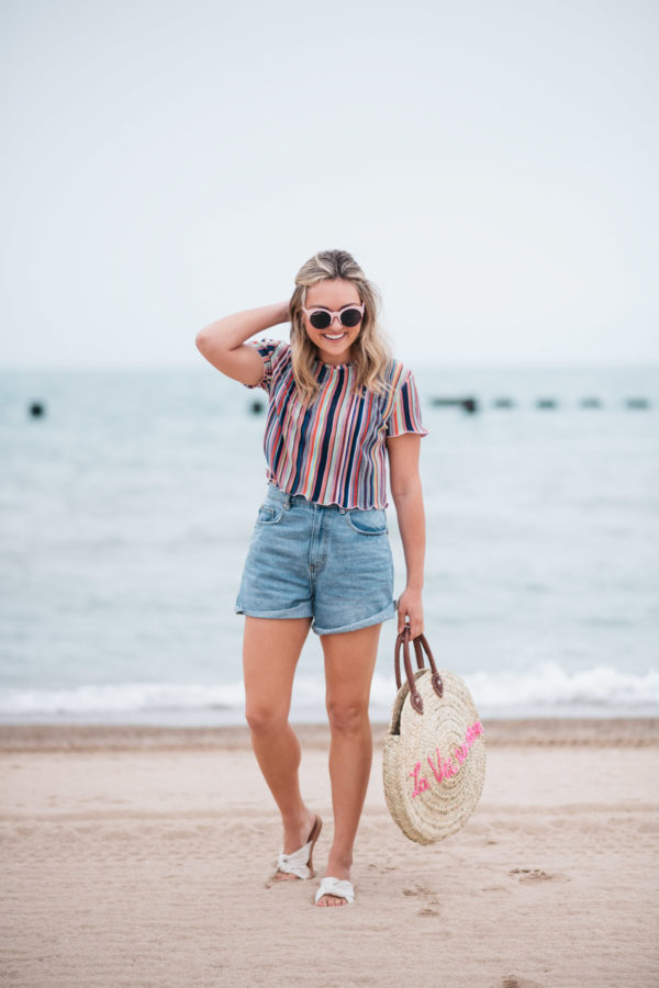 Fashion blogger Jessica Sturdy wearing pink sunglasses, a Bershka candy striped ruffled hem short sleeve cropped top, Cotton On high-rise shorts, and Soludos knotted slides with a White Elephant Designs embroidered tote by Lake Michigan in Chicago.