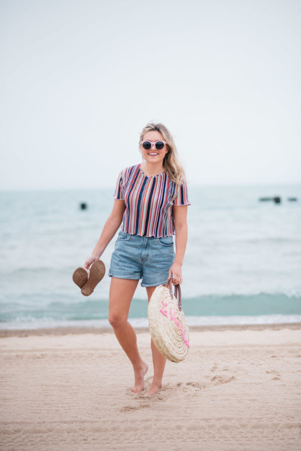 Jessica Sturdy with a La Vie en Rose embroidered straw tote, created in collaboration with White Elephant Designs, and wearing pink sunglasses, a Bershka striped cropped top and Cotton On high-rise flashback shorts with Soludos slide sandals at the North Avenue Beach in Chicago.