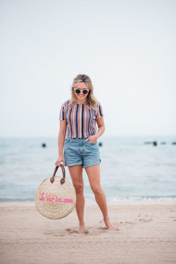 Jessica Sturdy with a La Vie en Rose embroidered straw tote, created in collaboration with White Elephant Designs, and wearing pink sunglasses, a Bershka striped cropped top and Cotton On high-rise flashback shorts with Soludos slide sandals at the North Avenue Beach in Chicago.