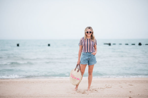 Jessica Sturdy with a La Vie en Rose embroidered straw tote, created in collaboration with White Elephant Designs, and wearing pink sunglasses, a Bershka striped cropped top and Cotton On high-rise flashback shorts with Soludos slide sandals at the North Avenue Beach in Chicago.
